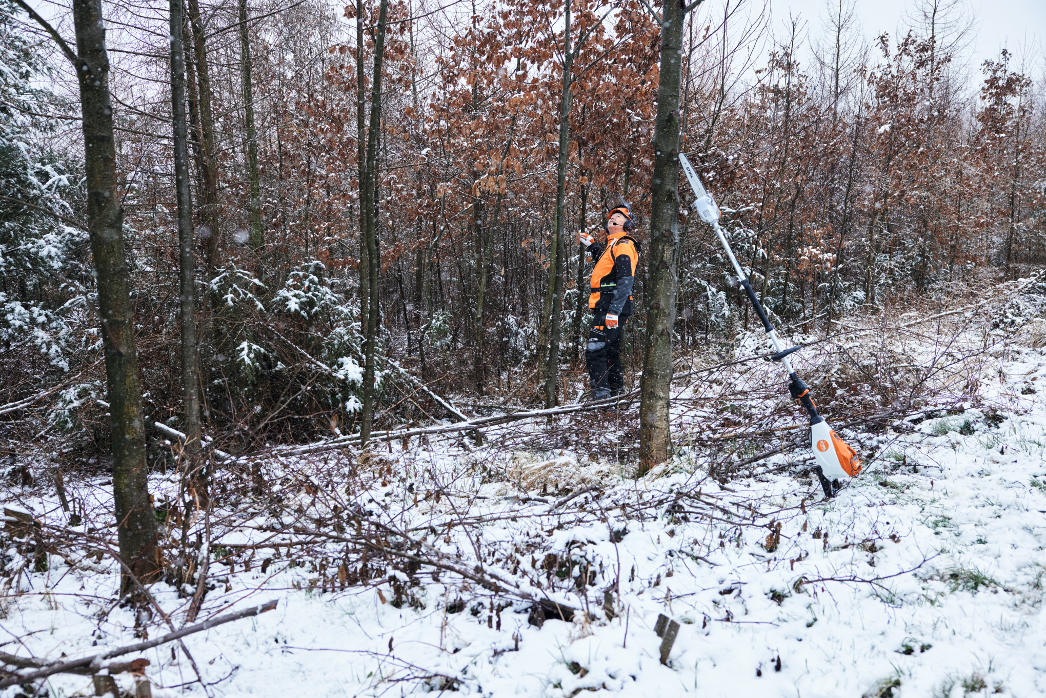 Un homme travaillant en équipement de protection dans une forêt enneigée, au premier plan une perche d’élagage à batterie STIHL HTA 150 posée contre un tronc d’arbre