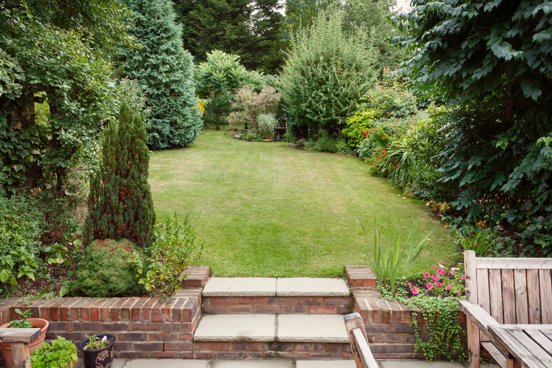 Petit jardin avec terrasse, mobilier de jardin et bacs à plantes au premier plan. En arrière plan, une pelouse avec des petits arbres et des arbustes.