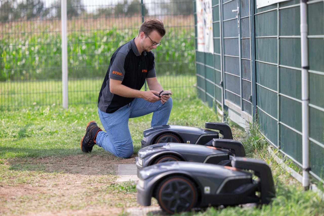 STIHL iMOW® Produktmanager Thilo Ehrentreich kniet neben drei Mährobotern mit einem Handy in der Hand