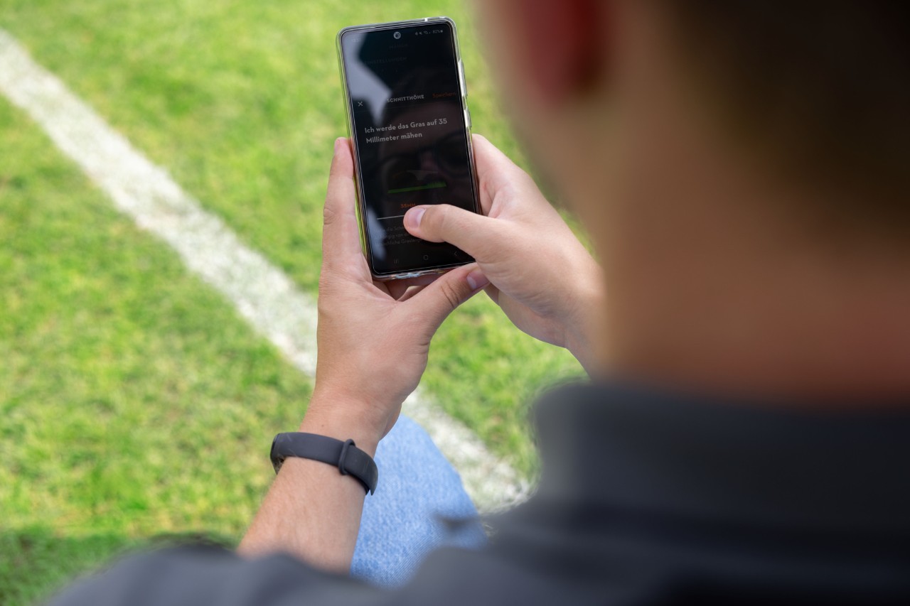 Eine Person auf einem Sportplatz hat die MY iMOW® App auf einem Handy geöffnet