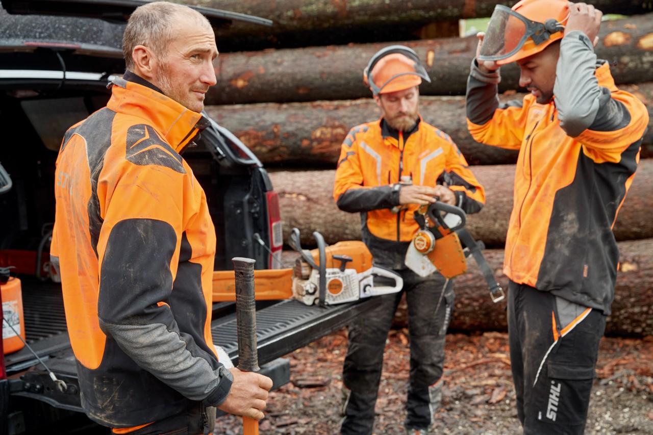 Drei Männer in STIHL Schutzkleidung vor einem Holzstapel bereiten sich auf die Arbeit mit einer STIHL Kettensäge vor