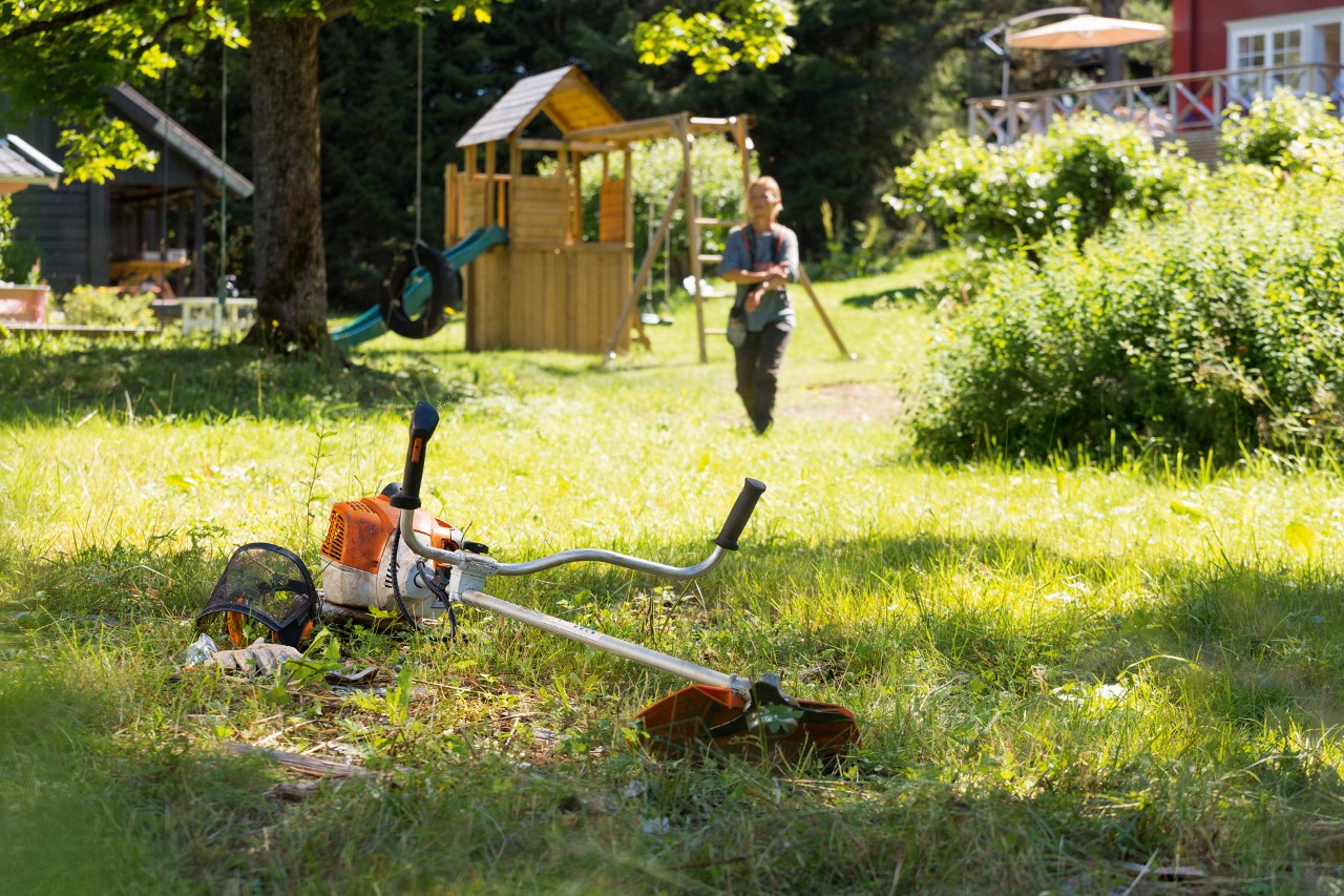 STIHL Motorsense FS 240 liegt im Gras, im Hintergrund bereitet ein Mann sich auf die Wartung vor