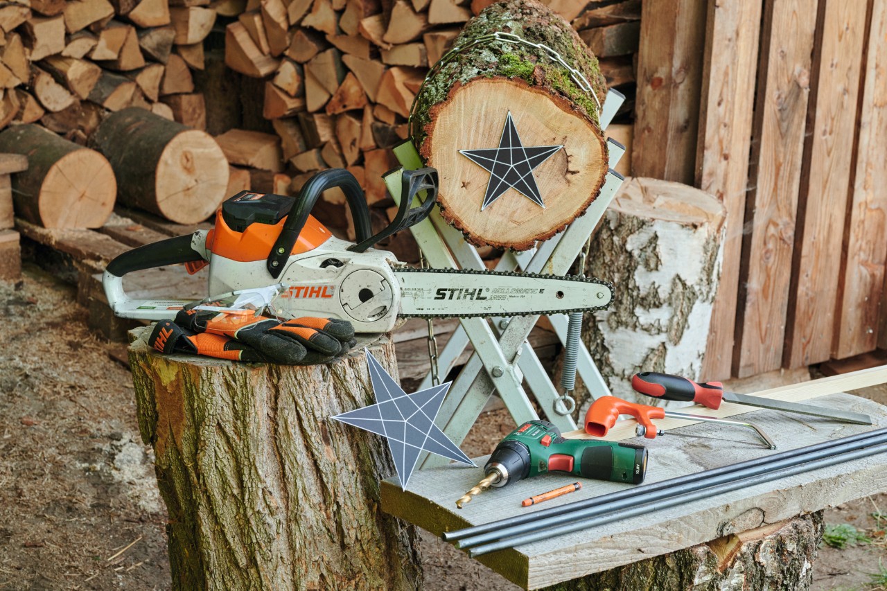 Vor einem Brennholzstapel liegen Schablone, Material und Werkzeug, um einen Holzstern selber zu machen