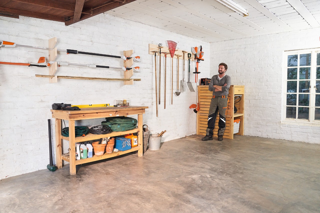 Mann in einer Werkstatt mit einem selbstgebauten Gartengerätehalter und verschiedenen STIHL Geräten, darunter Motorsensen und Laubrechen