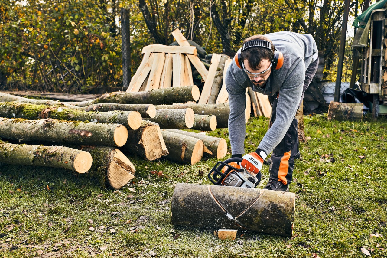 Ein Mann in Arbeitsschutzausrüstung sägt einen Baumstamm zurecht, um eine Gartenbank selber zu bauen 