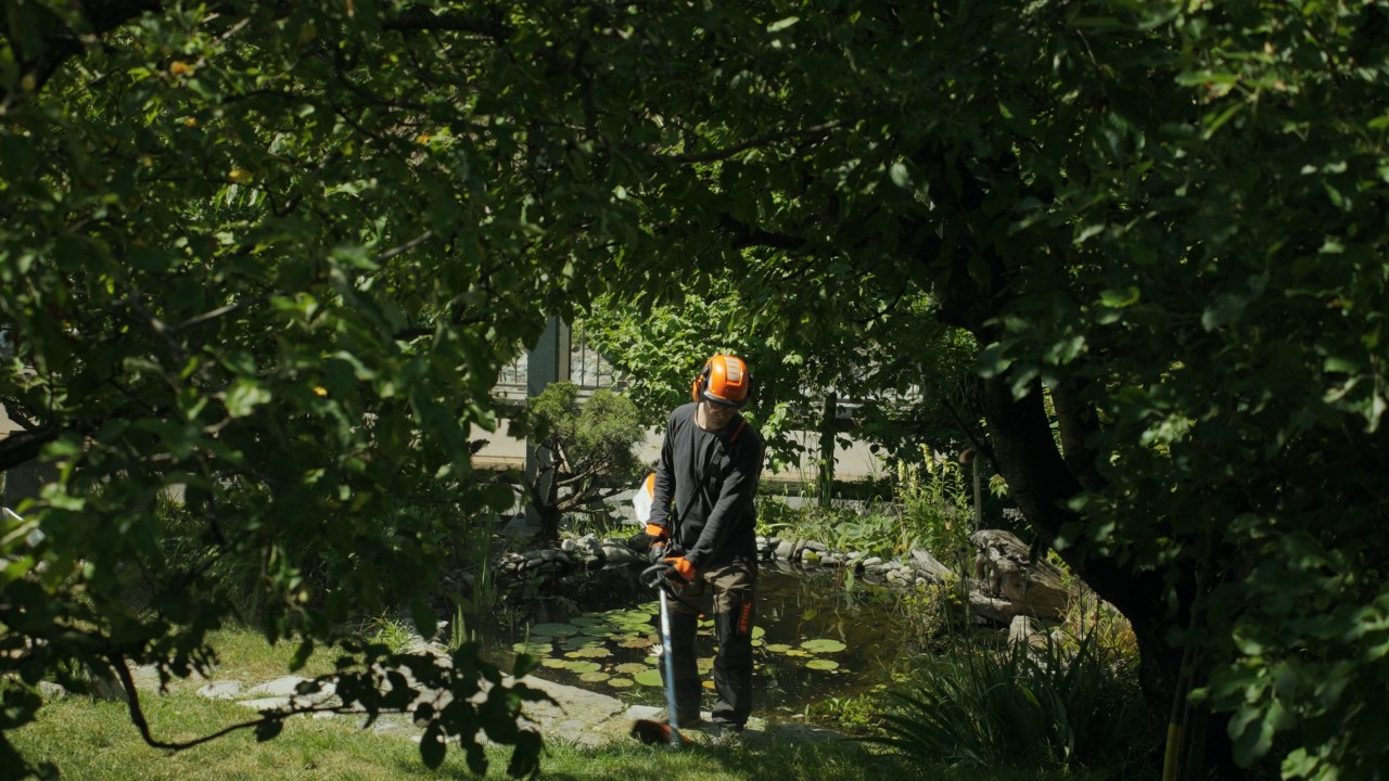 Achille arbeitet mit Freischneider im Garten und trägt Schutzkleidung von STIHL, im Hintergrund Gartenanlage mit Teich und grüner Vegetation, Real People. STIHL People. Kampagne