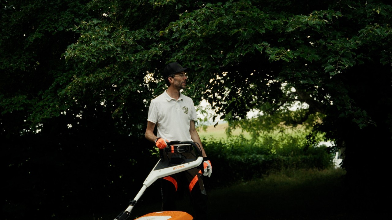 Patrick steht mit einem STIHL Rasenmäher im Garten und blickt auf die Rasenfläche, Real People. STIHL People. Kampagne