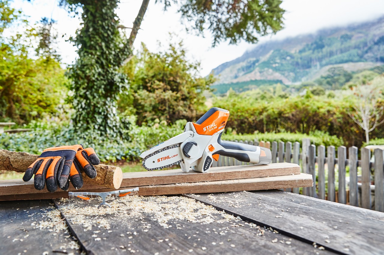 La scie de jardin GTA 26 est posée sur des planches sur une table de travail, à côté de gants de protection STIHL