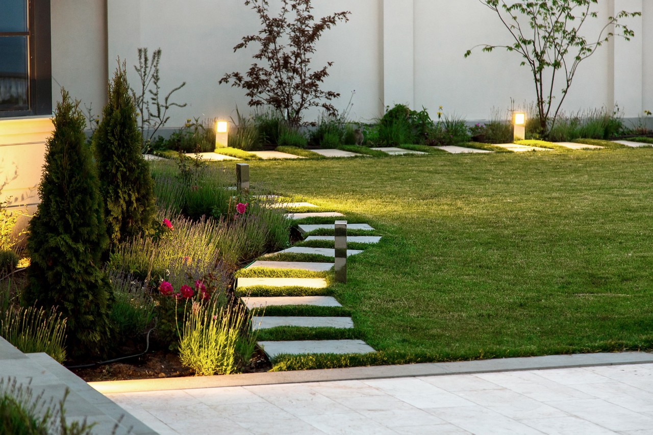 Petit jardin en soirée, parterre de fleurs, pelouse verte, chemin en dalles de marbre carrées, éclairage décoratif au sol.