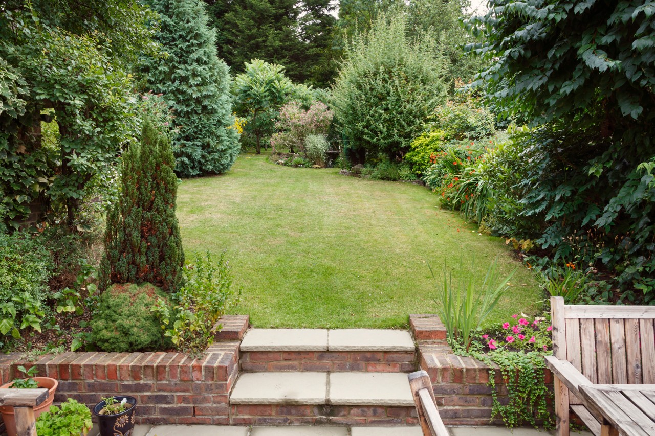 Petit jardin avec terrasse, mobilier de jardin et bacs à plantes au premier plan. En arrière plan, une pelouse avec des petits arbres et des arbustes.