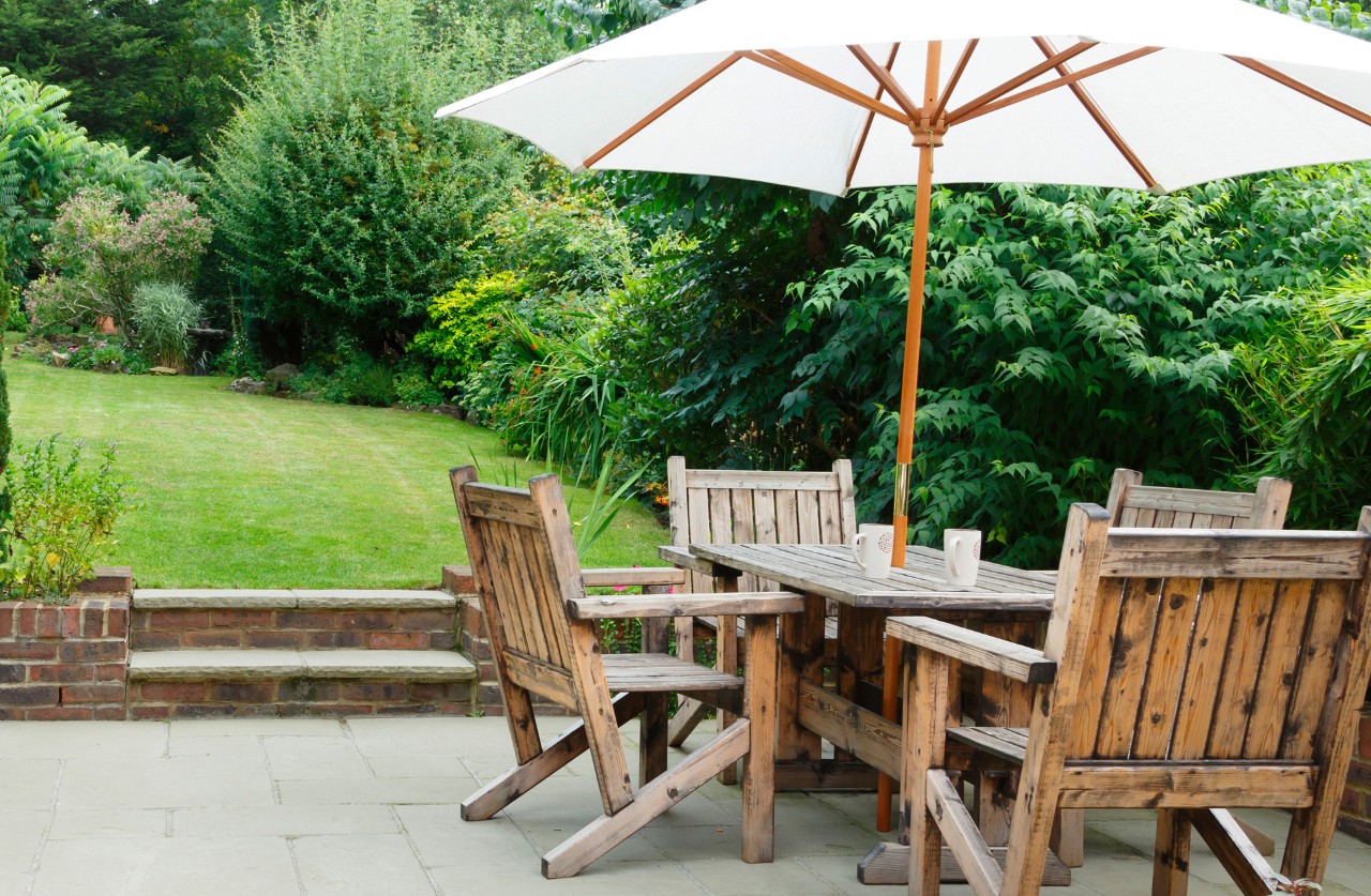 Jardin en contrebas avec pavés, mobilier de jardin au premier plan, pelouse avec arbustes et petits arbres à l’arrière-plan.