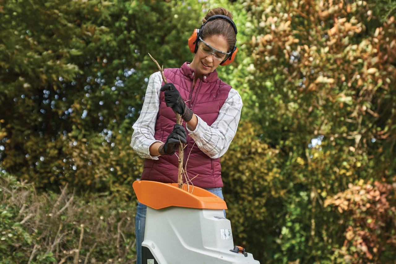 Femme portant des gants, des protections auditives et des lunettes en train d’insérer des branches dans un broyeur de jardin