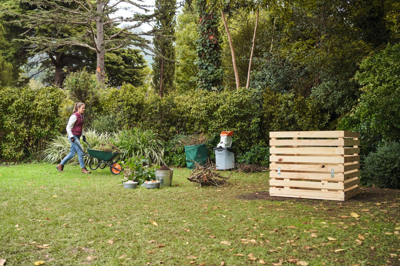 Une femme s’approche avec une brouette d’un bac à compost en bois qu’elle a construit elle-même, avec une haie dense et des arbres en arrière-plan 