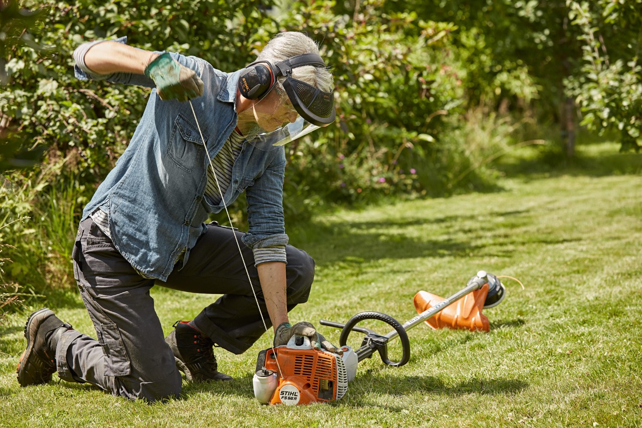 Une femme portant des protections auditive et faciale est en train de démarrer une débroussailleuse à essence STIHL FS 56 R sur un terrain vert