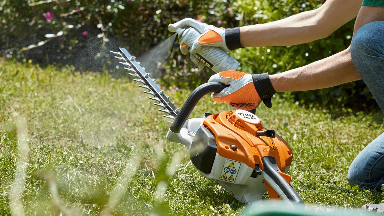 Un taille-haie thermique STIHL HS 82 T sur une pelouse avec une personne qui pulvérise du solvant pour résine sur l’outil de coupe