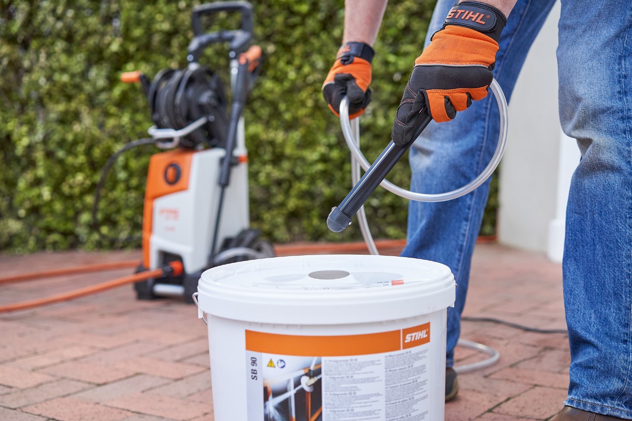 Un homme portant des gants de protection devant une haie de jardin utilisant l’abrasif de sablage STIHL SB 90 pour réaliser un sablage humide 