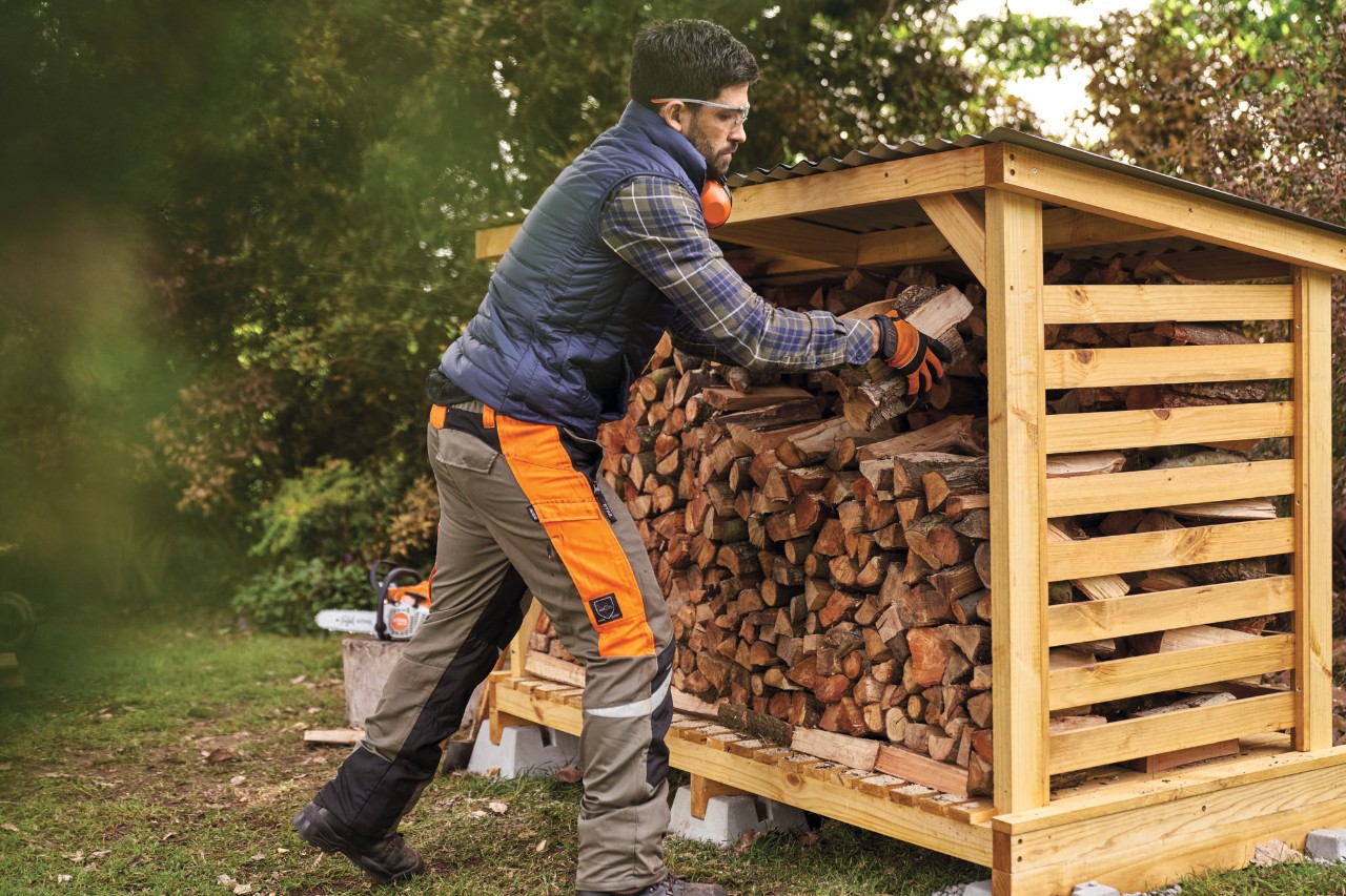 Un homme en vêtements de protection empile du bois de chauffage dans un abri à bois de chauffage qu’il a construit lui-même