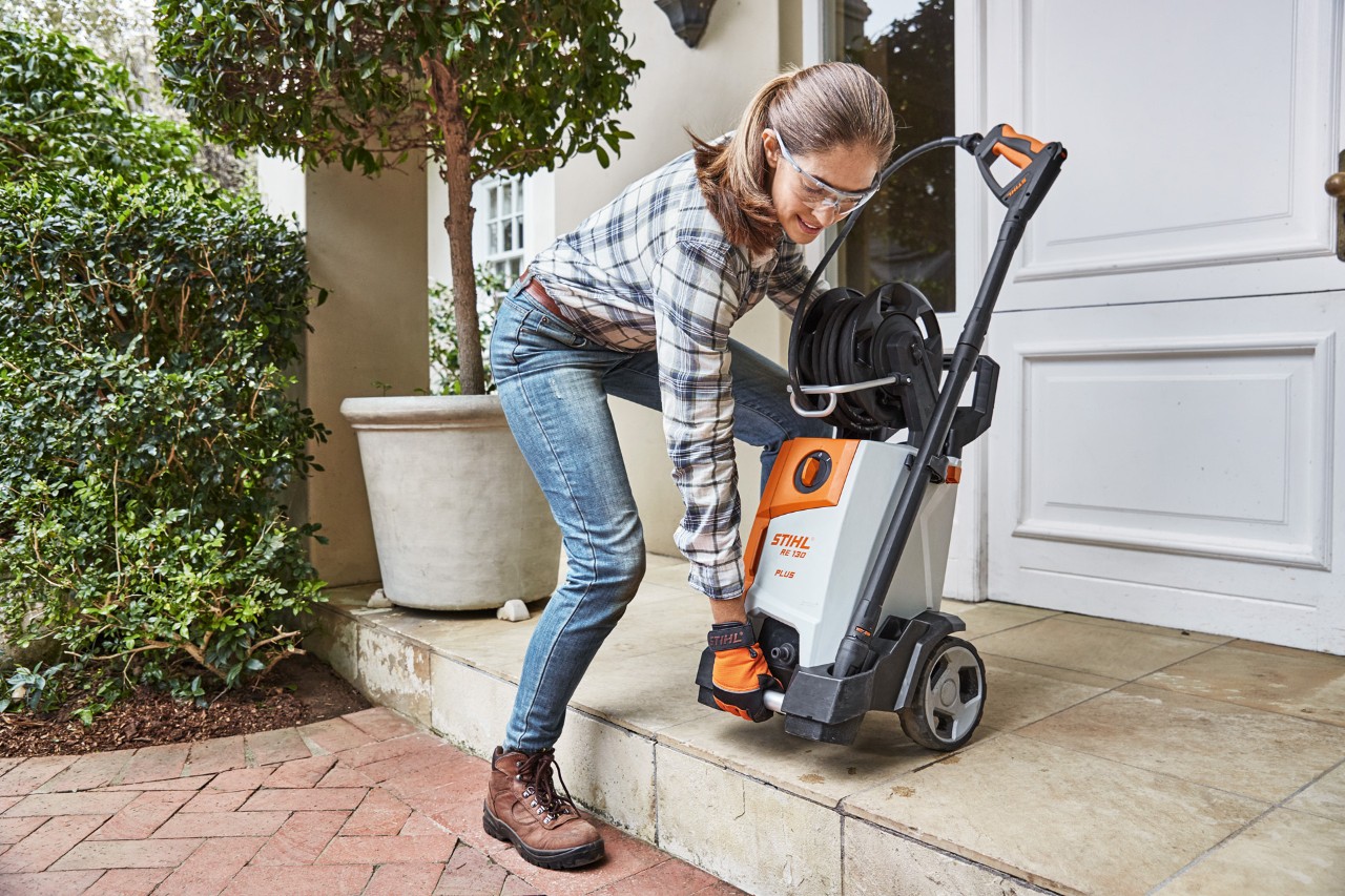 Une femme soulevant un nettoyeur haute pression STIHL RE 130 PlUS sur une allée en briques