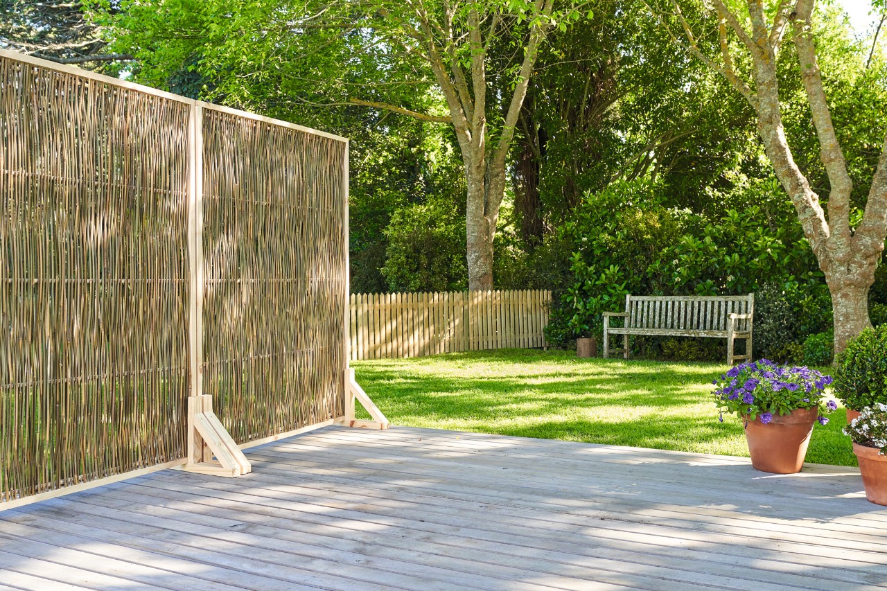 Pare-vue artisanal sur une terrasse, en arrière-plan une pelouse avec un banc devant des arbres 