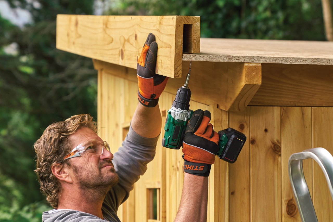 Comment construire un abri de jardin : homme montant un bord de toit pré-assemblé sur le toit d’un abri de jardin