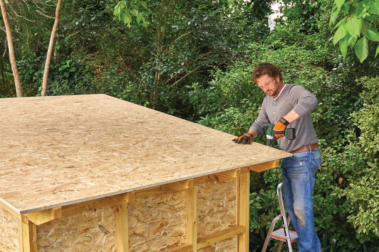 Comment construire un abri de jardin : homme sur une échelle vissant le panneau de toit en OSB sur un abri de jardin