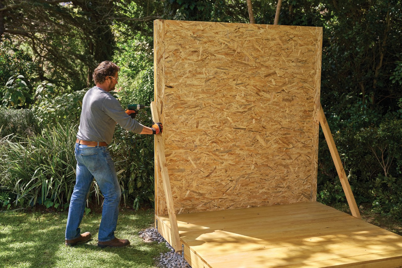 Un homme utilise des supports temporaires pour monter une paroi pré-assemblée sur la base d’un abri de jardin en bois