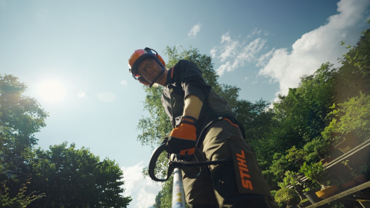 Achille travaille avec une débroussailleuse dans le jardin, porte un équipement de protection STIHL, avec de la végétation verte en arrière-plan, campagne Real People. STIHL People.