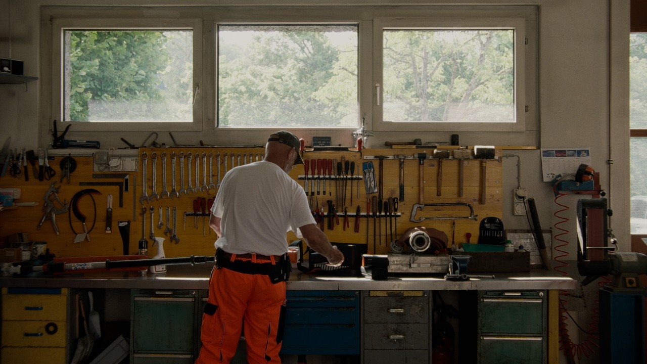 Lukas dans un atelier, vu de dos, tient une batterie STIHL et la retire d’un taille‑haie à batterie STIHL posé sur l’établi, campagne Real People. STIHL People.