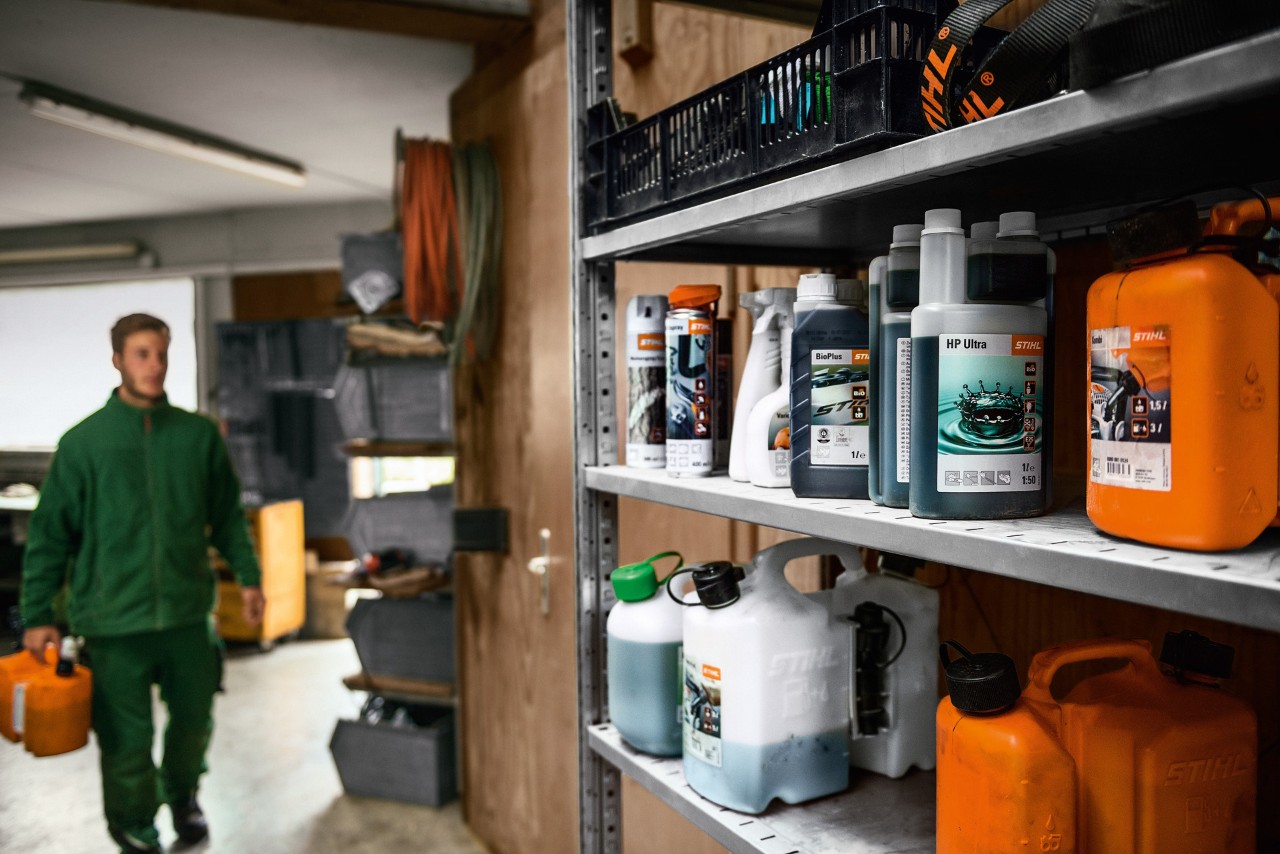Différents consommables STIHL sur une étagère, en arrière-plan un homme avec un bidon destiné au transport de matières dangereuses