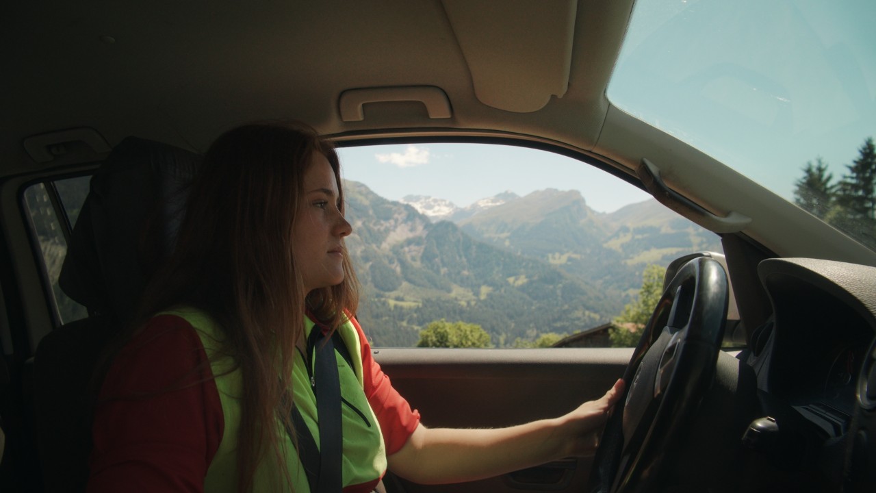 Bigna alla guida di un’auto, vista dall’interno con volante e paesaggio alpino sullo sfondo, Real People. STIHL People.