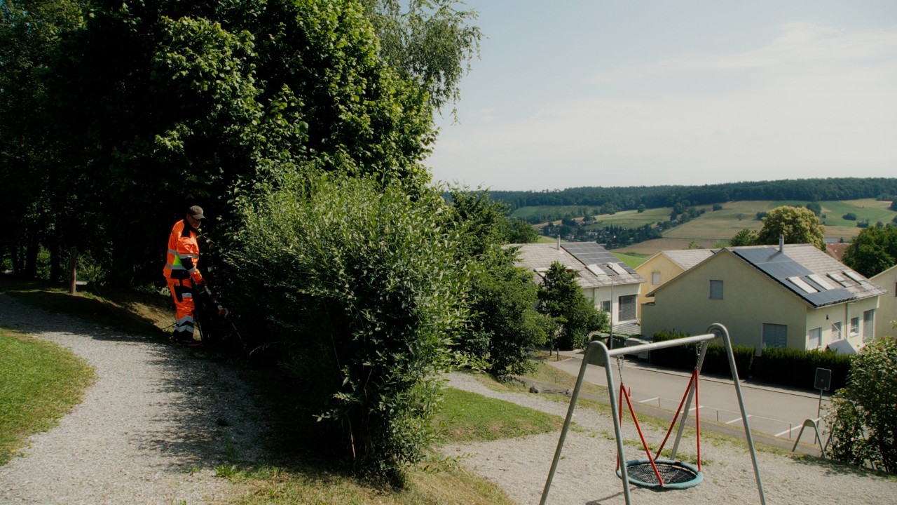 Lukas durante la manutenzione di una siepe con un tagliasiepi STIHL in un’area verde pubblica, con un parco giochi sullo sfondo, campagna Real People. STIHL People.
