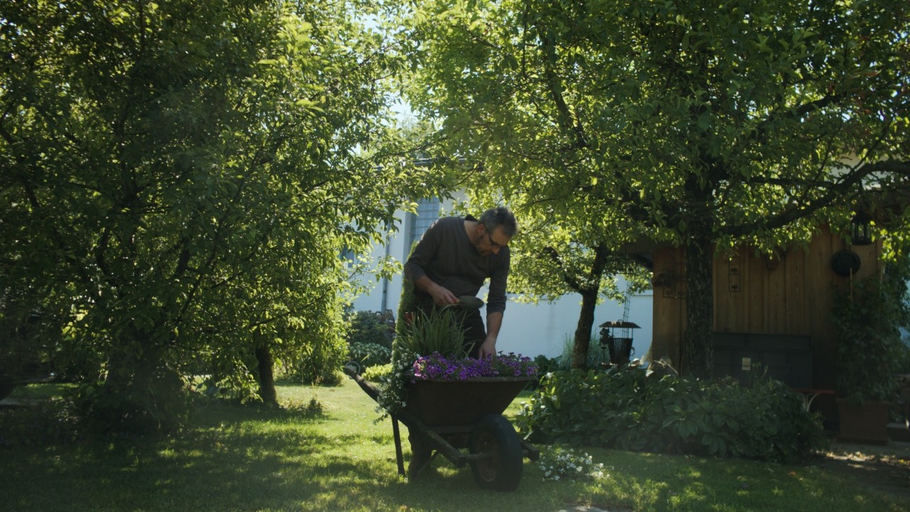 Rolf lavora in giardino con una pala sui fiori in una carriola, prato verde e piante sullo sfondo, campagna Real People. STIHL People.