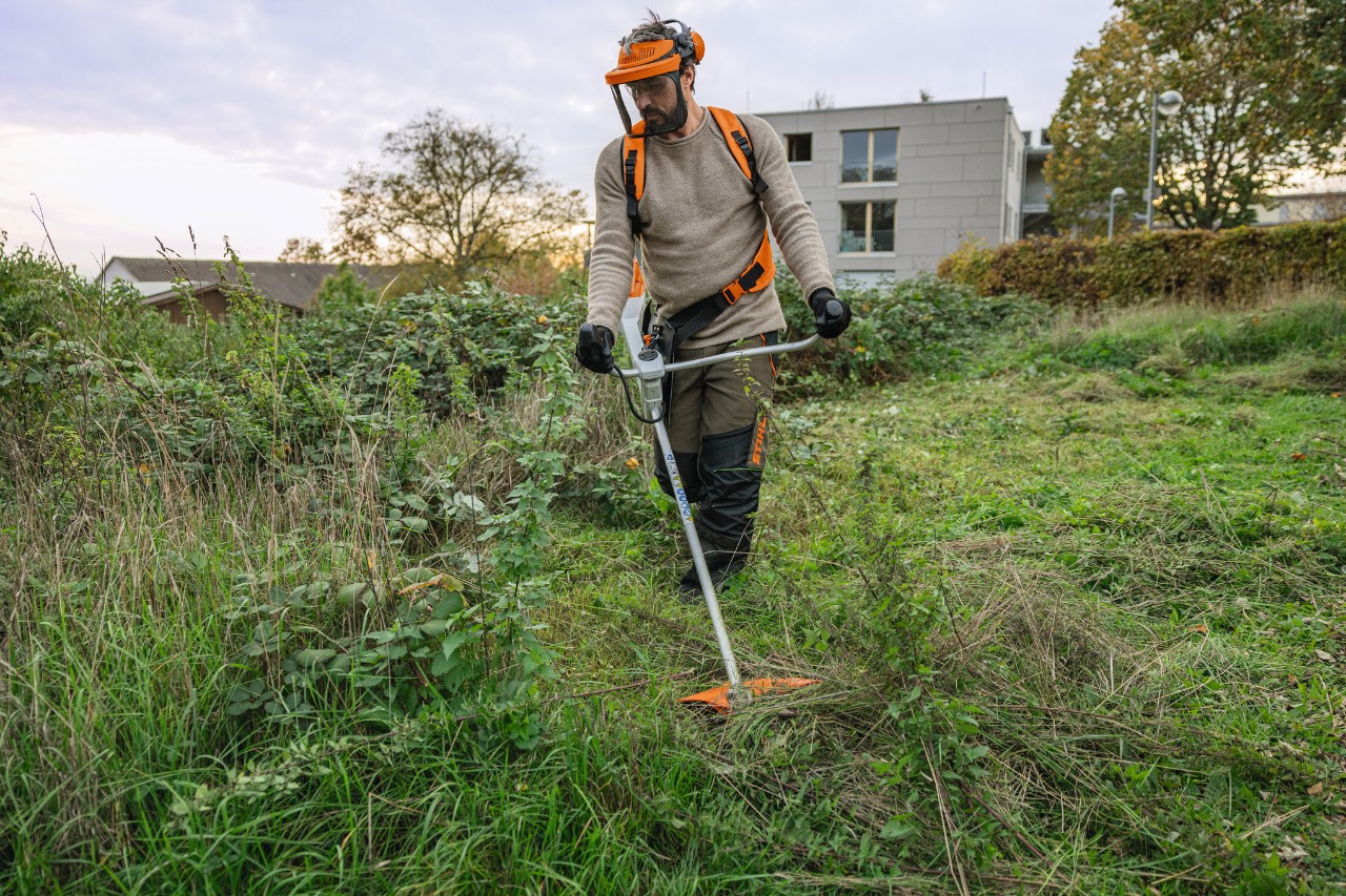 Un uomo che indossa l'equipaggiamento protettivo STIHL falcia un'area verde selvaggiamente ricoperta di vegetazione con un decespugliatore a batteria STIHL FSA 200