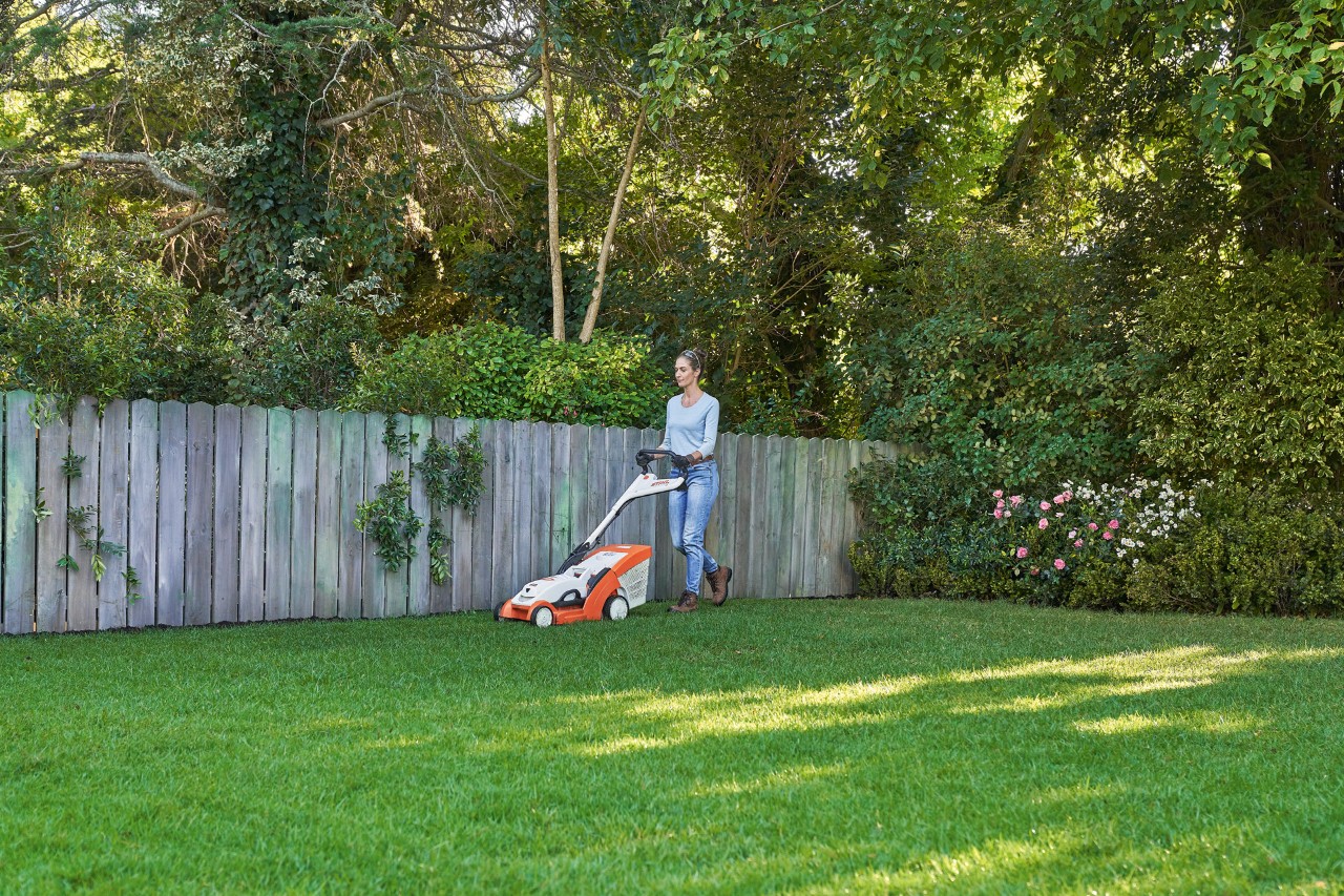 Donna che falcia un prato verde davanti a una staccionata di legno con il tosaerba a batteria STIHL RMA 339 C.
