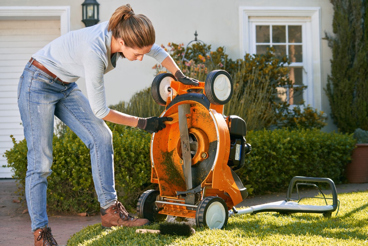 Donna che pulisce la parte inferiore di un tosaerba a benzina STIHL RM 448 T-C con una spatola