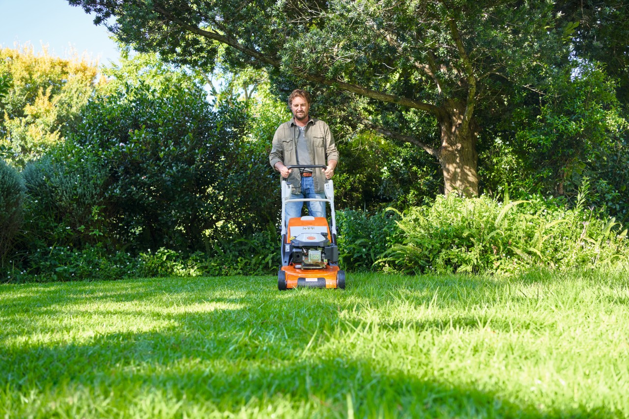 Un uomo falcia un prato semi-ombreggiato con un tosaerba STIHL, sullo sfondo si vedono cespugli e alberi