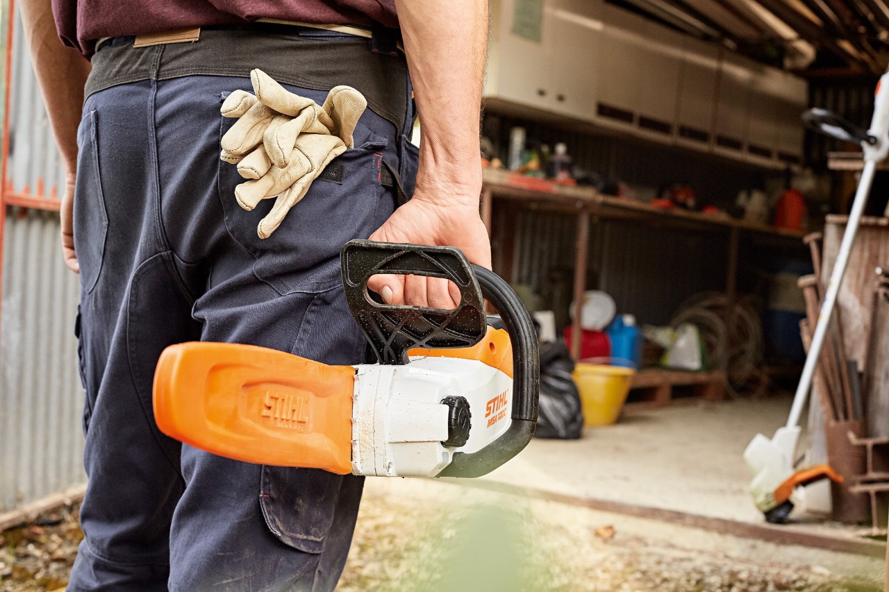Uomo che sorregge una motosega senza fili STIHL con spranga di guida, sullo sfondo è visibile un decespugliatore 